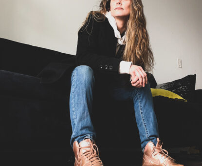 Monaya M. MaGaurn, lifestyle physicist, sitting on a black couch in blue jeans, gold sneakers, with a black jacket, white cuffs and collar, with long hair.
