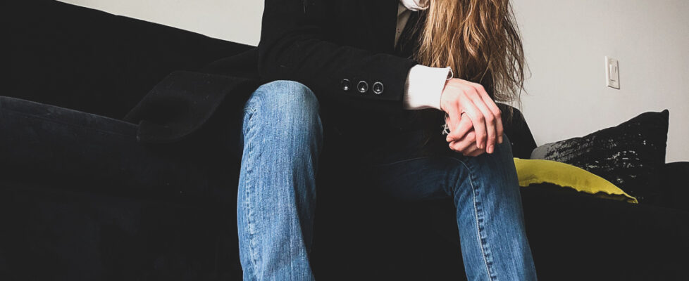 Monaya M. MaGaurn, lifestyle physicist, sitting on a black couch in blue jeans, gold sneakers, with a black jacket, white cuffs and collar, with long hair.