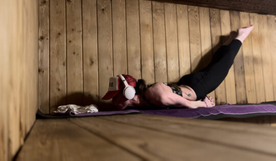 Monaya M. MaGaurn, lifestyle physicist with Agency World Resources WTR, lying on a yoga mat inside a wooden sauna. She wears a red cap, white over-ear headphones, black leggings, and a patterned sports bra. She is in a supported backbend with hands clasped behind her, chest lifted, and legs extended straight.