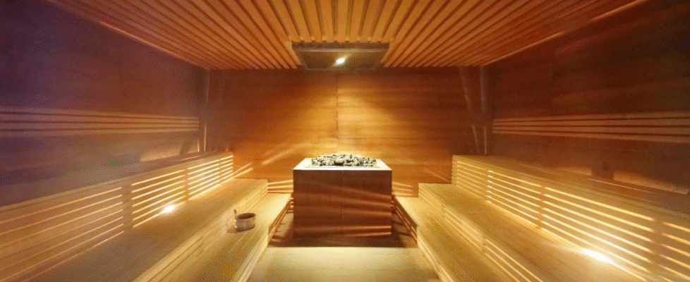 Warmly lit wooden sauna interior with symmetrical tiered benches on both sides and a central stone heater. Soft light glows through slats, creating a calm, inviting atmosphere for relaxation and heat therapy.